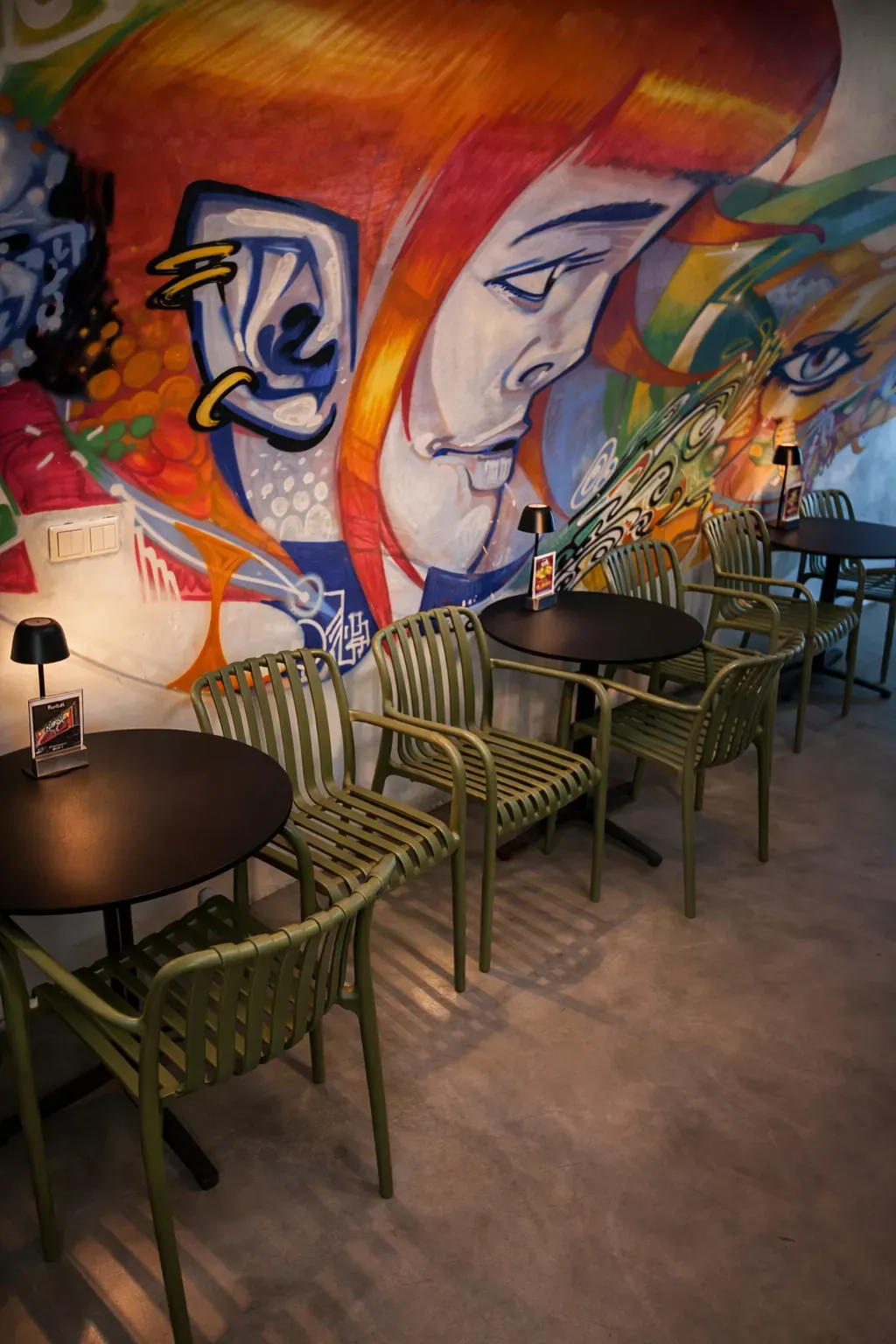 Sedute di un caffè al chiuso con sedie verdi e tavoli neri davanti a un murale colorato con un volto.
